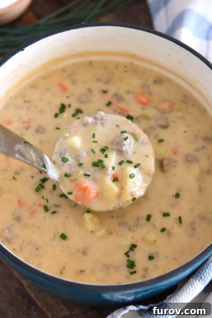 A ladle holding a scoop of creamy cheesy soup, featuring potatoes, hamburger, and carrots, garnished with fresh chives, ready to be served.