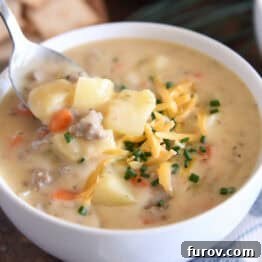 A white bowl with a spoon scooping creamy cheesy soup. The soup contains cooked potatoes, ground beef, and carrots, and is topped with shredded cheddar cheese and fresh chopped chives.