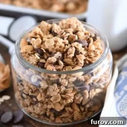 Shallow glass jar filled with peanut butter granola with roasted peanuts and dark chocolate chunks with spoonful of peanut butter to the side.