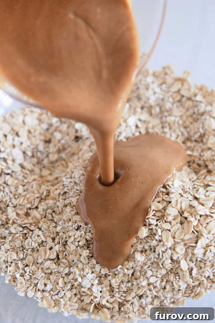 Pouring the warm peanut butter syrup mixture over dry oats in a large glass bowl, ensuring even coating.