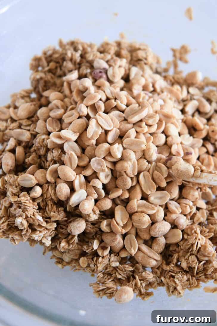 Roasted peanuts being added on top of the wet peanut butter granola mixture in a glass bowl, ready to be tossed.