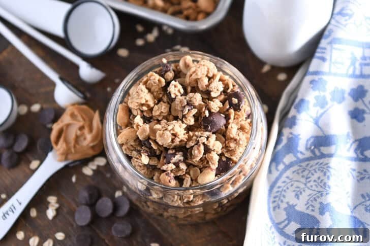 Top down view of a shallow glass jar filled with homemade peanut butter dark chocolate chunk granola with roasted peanuts and dark chocolate chunks. A spoonful of peanut butter is placed beside it.