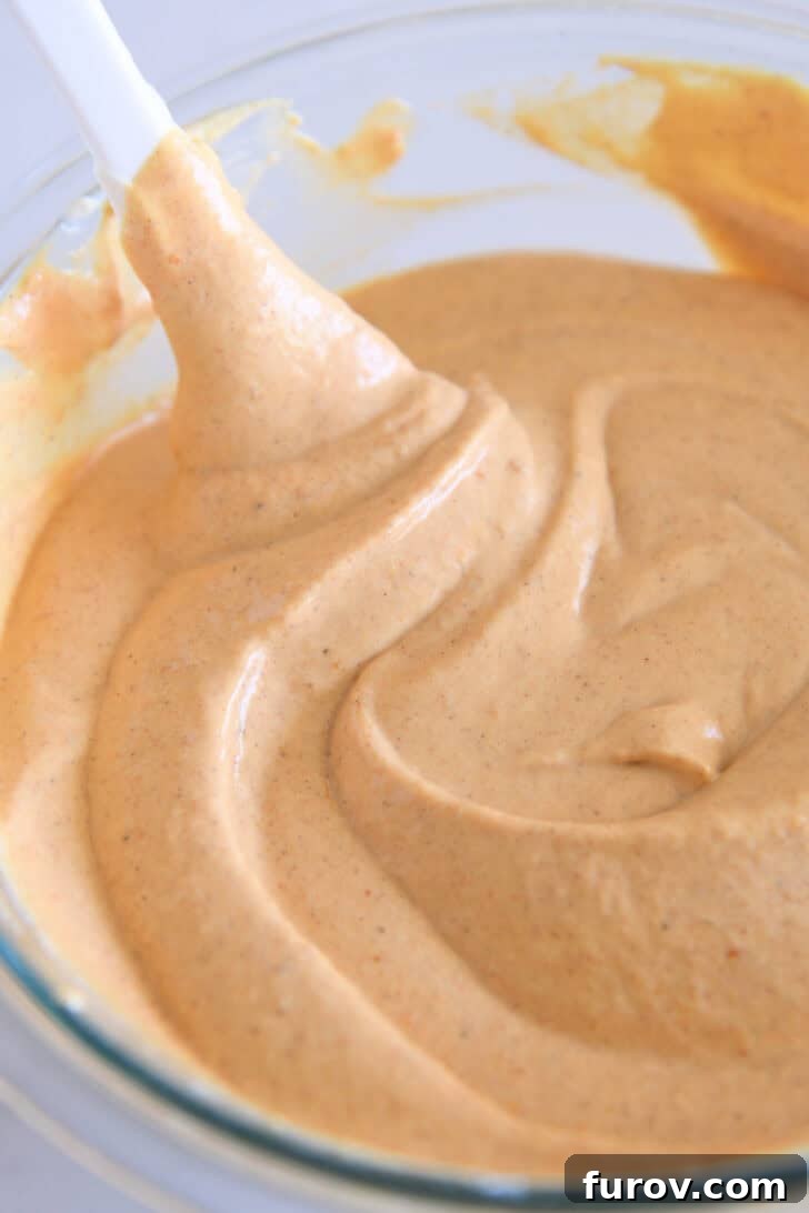 Golden Pumpkin Swirl Cheesecake 6 A vibrant pumpkin cheesecake batter in a glass bowl, with a white spatula demonstrating its smooth, well-combined texture.