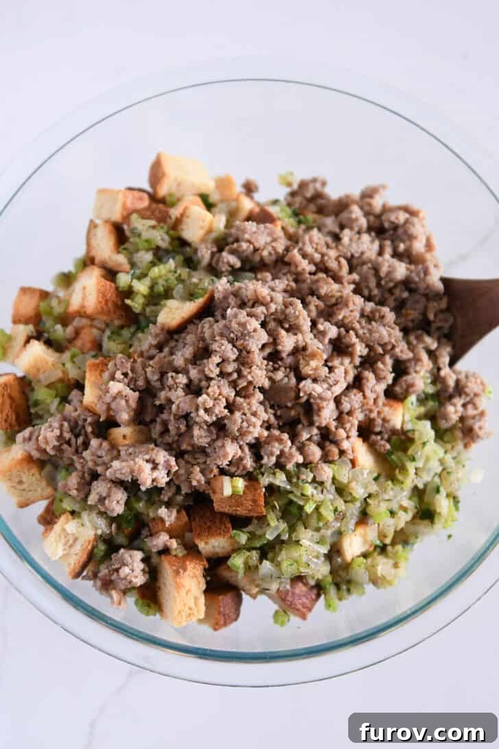 Large glass bowl containing toasted bread cubes, cooked vegetables, and crumbled sausage, ready for broth.