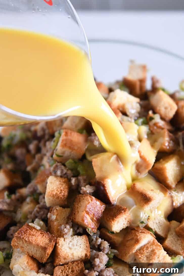 Hand pouring blended eggs and chicken broth over a mixture of dried bread cubes, cooked sausage, and sautéed herbs.