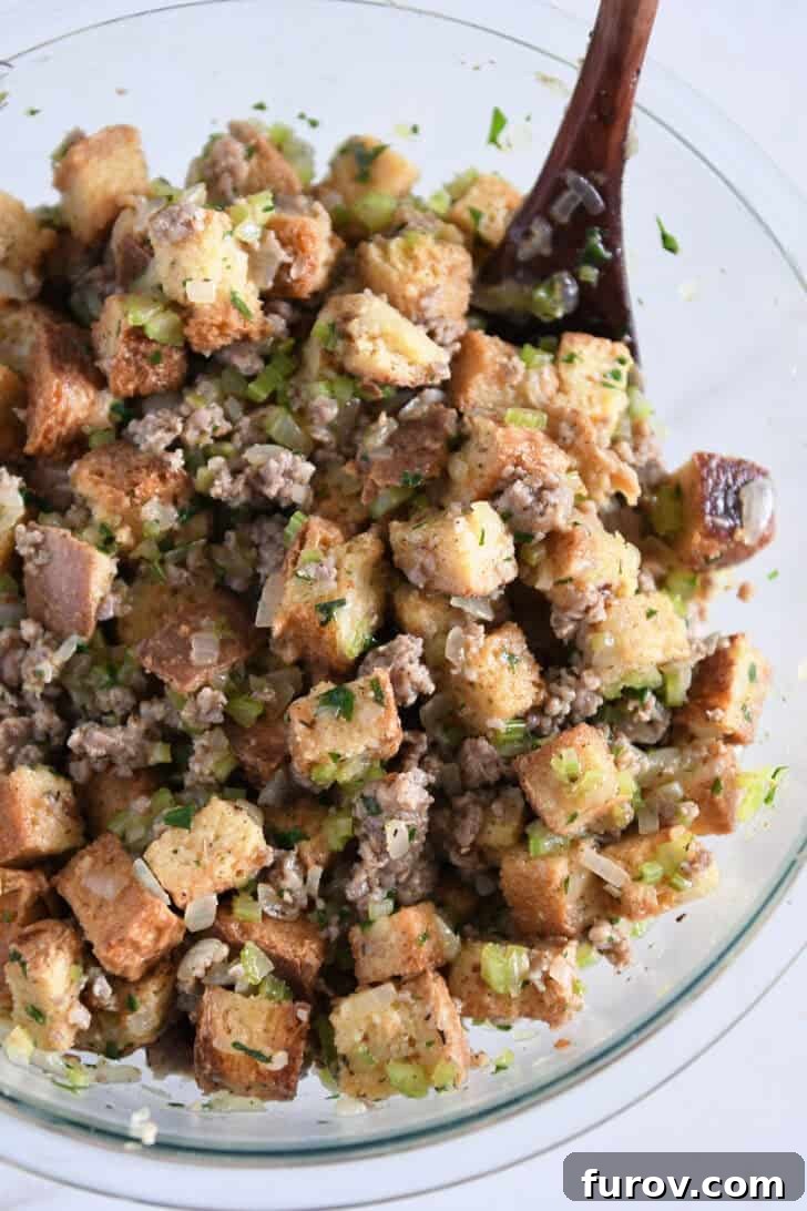 Glass bowl filled with toasted bread cubes, cooked sausage, sautéed vegetables, thoroughly saturated with broth and eggs, ready for baking.