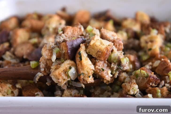 A serving spoon lifting a generous portion of baked sausage and herb stuffing from a pan, showing crispy top and moist interior.