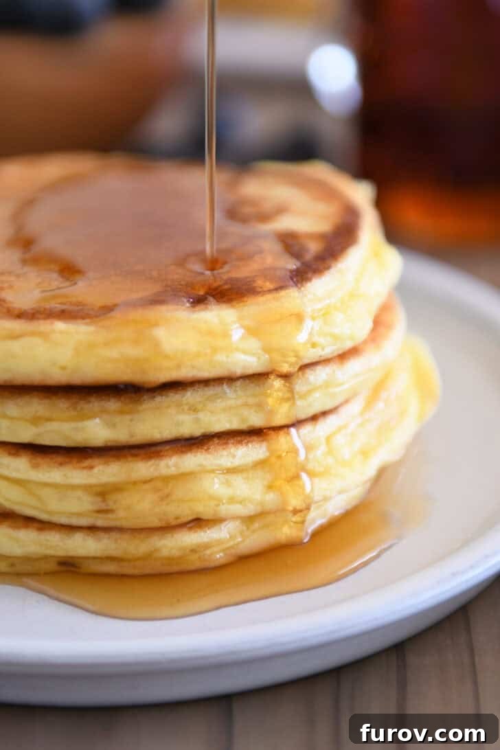 Drizzling pure maple syrup over a stack of fluffy cottage cheese pancakes on a gray plate.