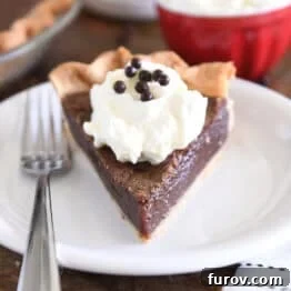 Triangle slice of chocolate chess pie on white plate with fork to the side and a dollop of whipped cream on top of pie topped with chocolate crisp pearls.