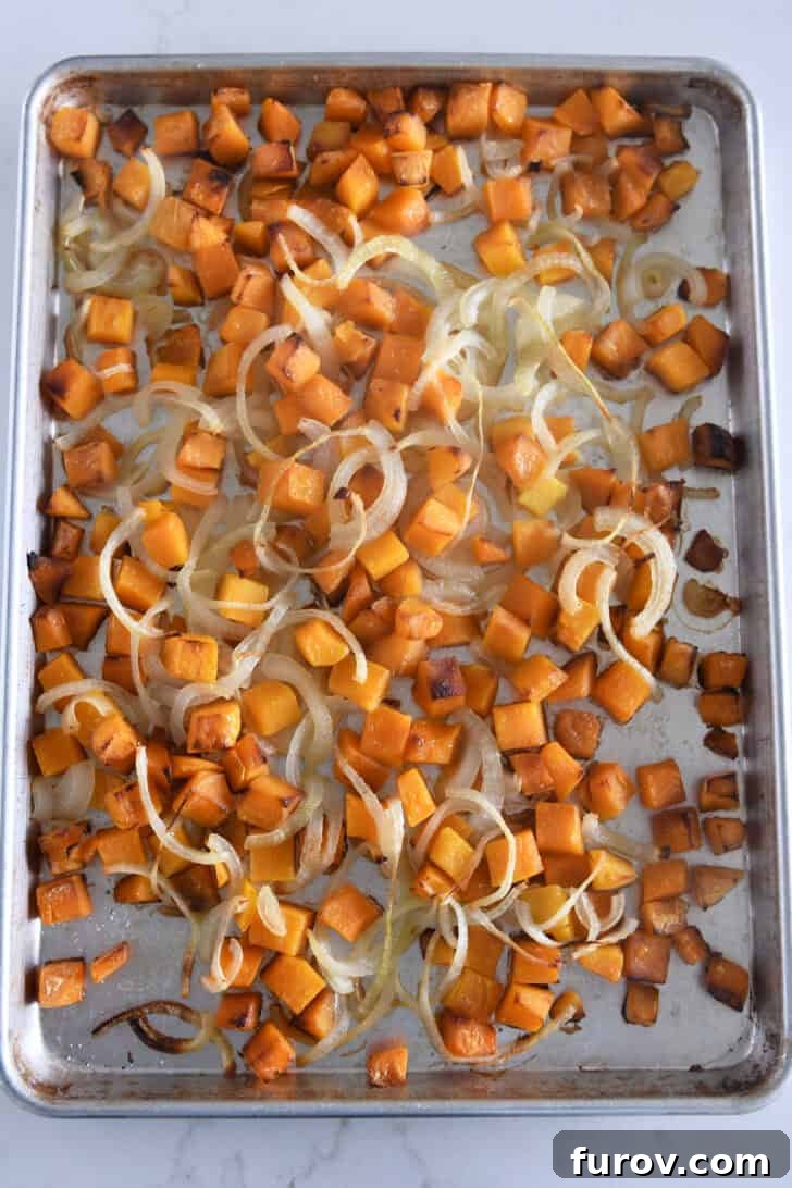Half sheet pan with cooked butternut squash pieces and slivered onions, golden and caramelized from roasting.