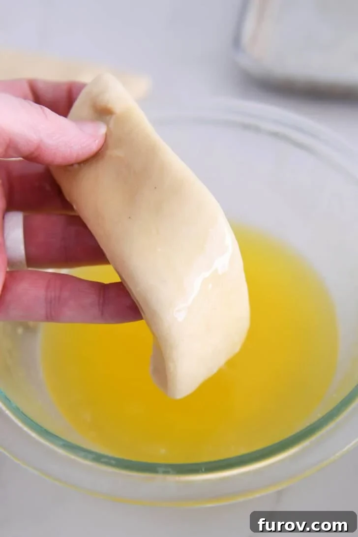 A piece of dough being thoroughly dipped into a bowl of melted butter, ensuring full saturation.