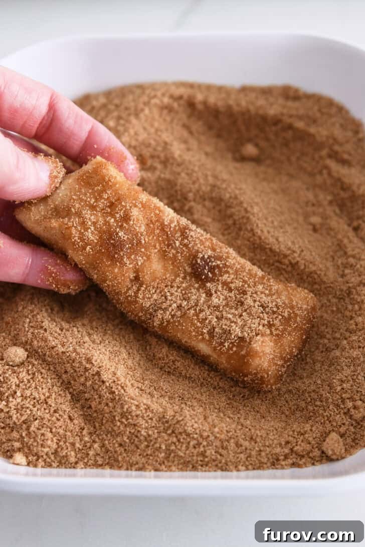 A butter-coated piece of dough being dipped into a bowl of cinnamon and sugar mixture, receiving its sweet topping.