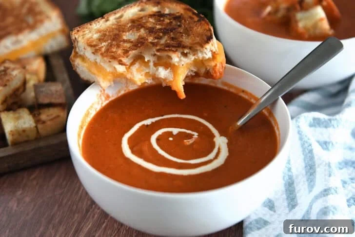 White bowl with homemade tomato soup, swirls of heavy cream, and half a grilled cheese sandwich balanced on side of bowl.