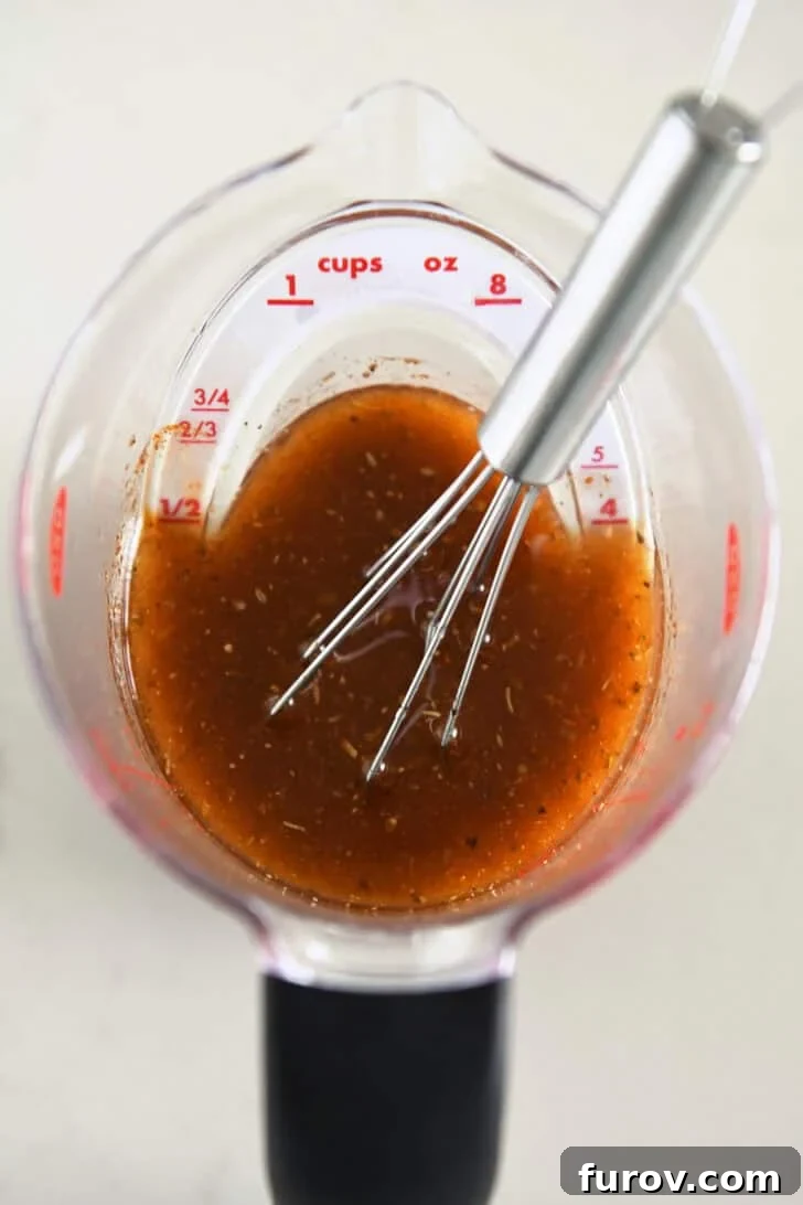 A clear liquid measuring cup filled with a vibrant orange-red chicken marinade, with a small whisk resting inside, ready to flavor the chicken.