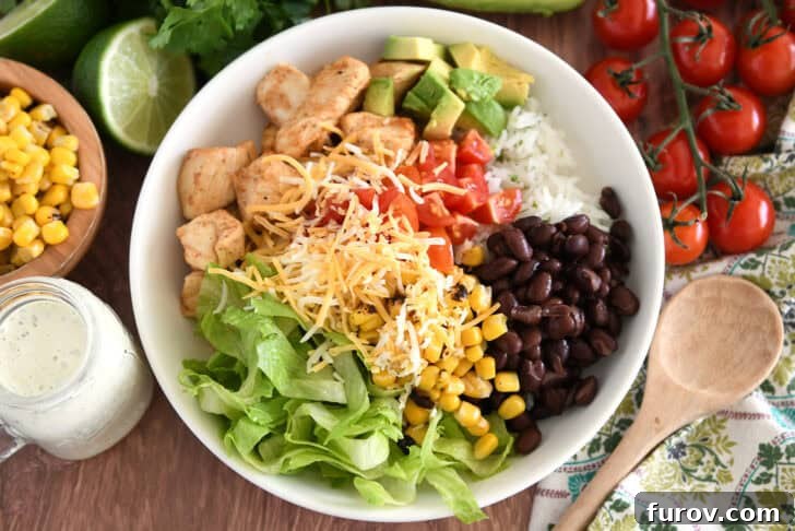 A fully loaded white bowl with cilantro lime rice, chicken, black beans, lettuce, roasted corn, fresh chopped avocados, diced tomatoes, and a sprinkle of shredded cheese.