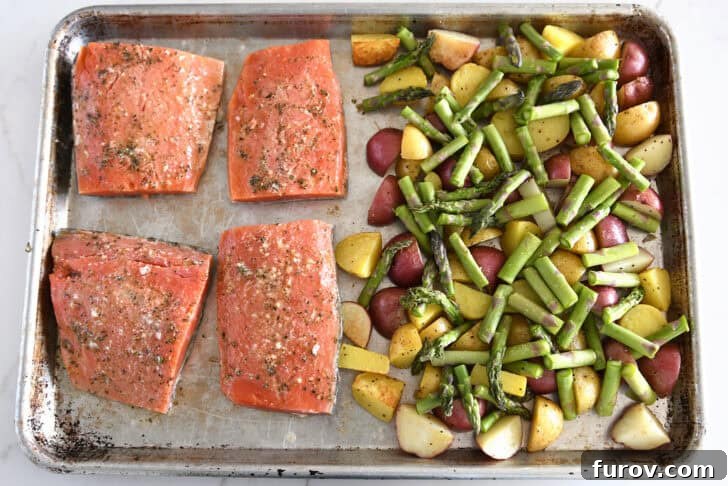 Top-down view of a perfectly arranged sheet pan featuring vibrant asparagus, roasted potatoes, and four seasoned salmon fillets, all ready for the oven.