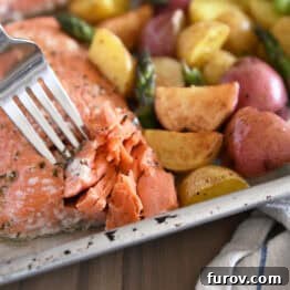 Fork flaking a filet of perfectly baked lemon garlic salmon with roasted potatoes and asparagus on a sheet pan, showcasing a complete and appetizing meal.