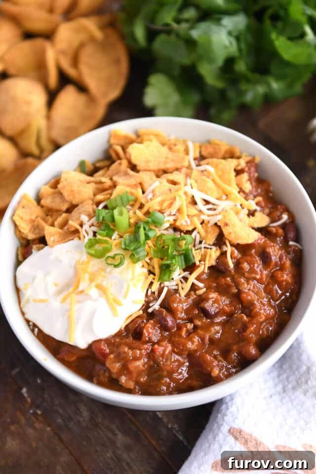 A rich, red chili with beans and ground beef in a rustic gray bowl, generously topped with creamy sour cream, shredded cheese, fresh green onions, and crispy crushed corn chips, ready to be enjoyed.