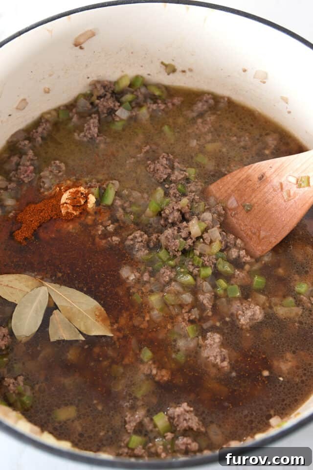 A cast iron pot containing perfectly cooked ground beef, seasoned with vibrant chili powder, fragrant bay leaves, and diced celery, actively simmering to develop deep flavors.