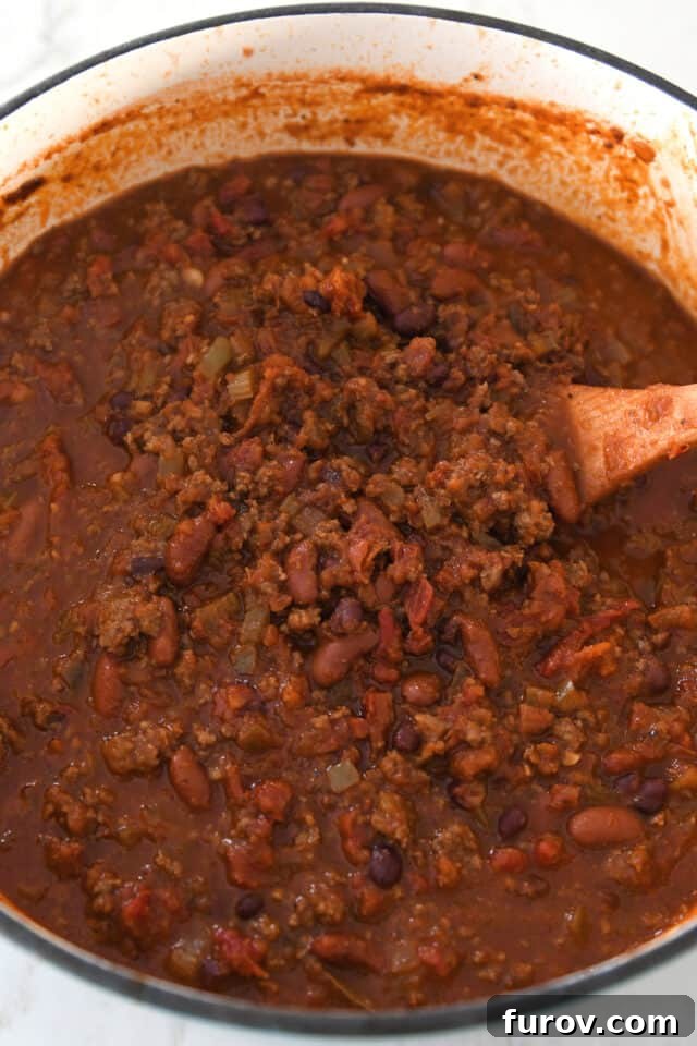 A large cast iron pot holds a simmering, cooked red chili, brimming with tender ground beef and a mix of flavorful beans, showcasing its rich, thick texture.