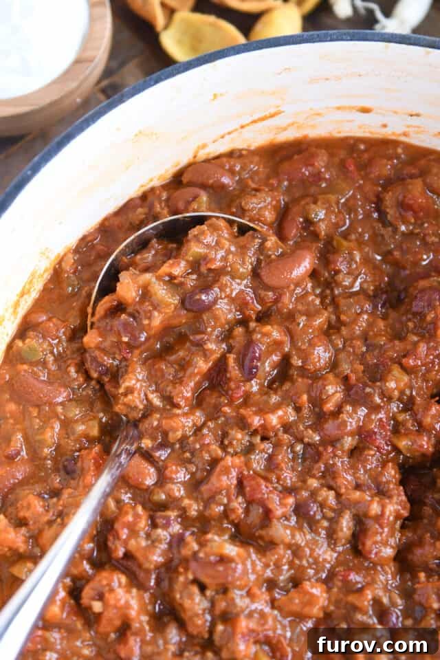 A pristine white cast iron pot filled with savory red chili, rich with ground beef and beans, with a ladle ready to serve, highlighting its inviting texture and warmth.