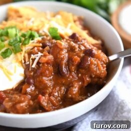 A close-up of red chili with a spoonful lifted from a gray bowl, garnished with creamy sour cream, vibrant green onions, and crunchy crushed corn chips.