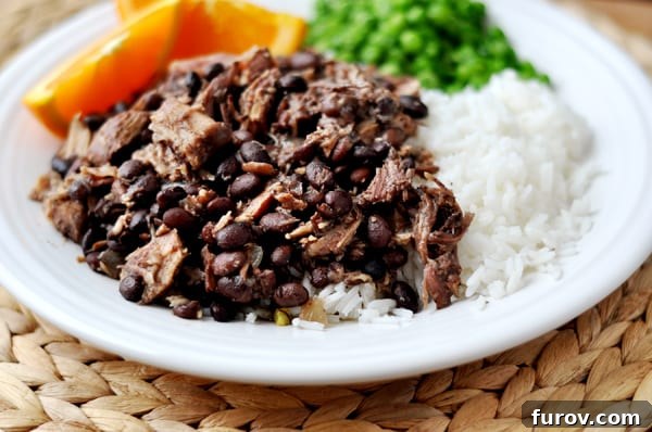 Shredded pork and black beans next to cooked white rice, peas, and orange slices on a white plate, showcasing a complete meal.