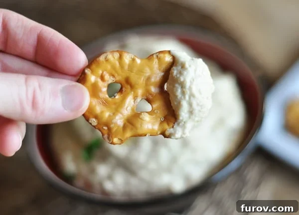 Someone holding a pretzel dipped in creamy homemade hummus over a bowl of the dip, highlighting a popular serving suggestion.