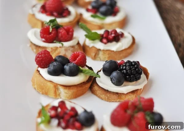 Top-down view of a white tray displaying an assortment of sweet berry bruschetta, adorned with pomegranate, raspberries, blackberries, and blueberries.