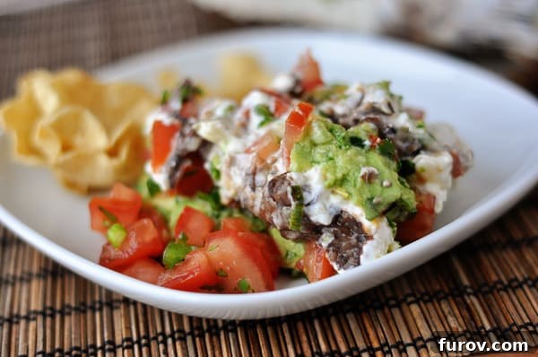 White plate with a big serving of 7-layer dip and tortilla chips on the side.