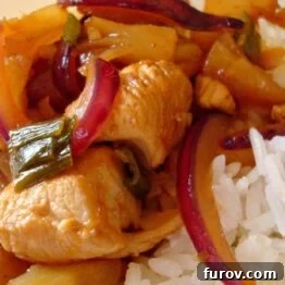 Close-up of sweet and sour chicken with pineapple and red onion, glistening with sauce, served over rice.