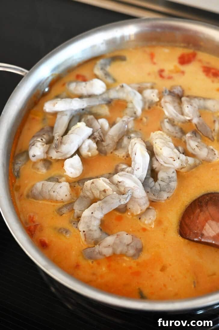Raw shrimp going into a metal pot with a rich, bubbling tomato curry sauce, ready to be cooked.