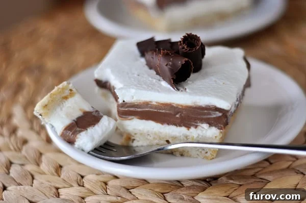 A delectable piece of Robert Redford Dessert on a white plate, showcasing its distinct layers: a golden shortbread crust, a smooth pudding middle, and a cloud of cool whip topping adorned with delicate chocolate curls.