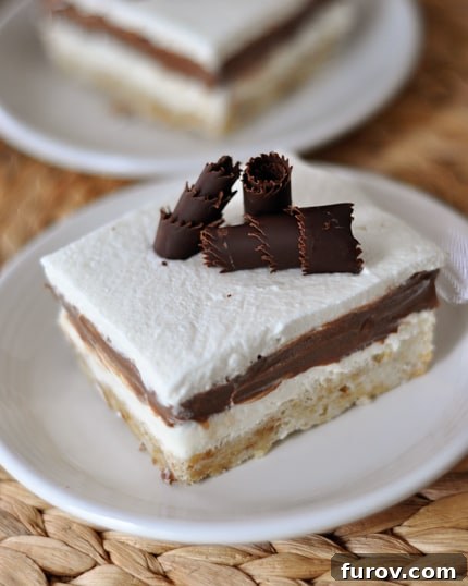 A close-up shot of a single slice of Robert Redford Dessert, highlighting the delicate balance of its shortbread crust, thick chocolate pudding, and generous topping of sweetened whipped cream and chocolate curls.