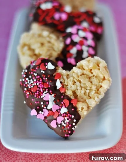 Chocolate-Dipped Krispie Hearts 3 White tray showcasing an array of heart-shaped Rice Krispie treats, each half-dipped in luscious chocolate and generously adorned with vibrant sprinkles.