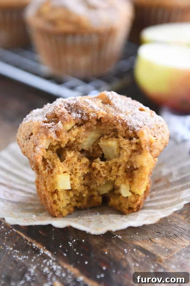 A close-up of a pumpkin apple spice muffin with a bite taken out, revealing the fluffy interior and apple chunks, resting in a brown paper liner.