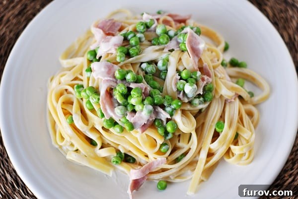 Savory Ham and Sweet Pea Fettuccine 3 Top view of a white plate with creamy fettuccine topped with chopped ham and green peas.