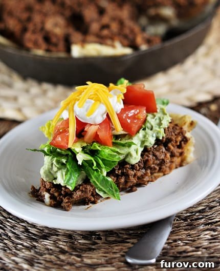 A perfectly portioned slice of Skillet Taco Pie on a white plate, generously topped with fresh lettuce, diced tomatoes, sour cream, and shredded cheese. The golden-brown masa harina crust is visible at the bottom.