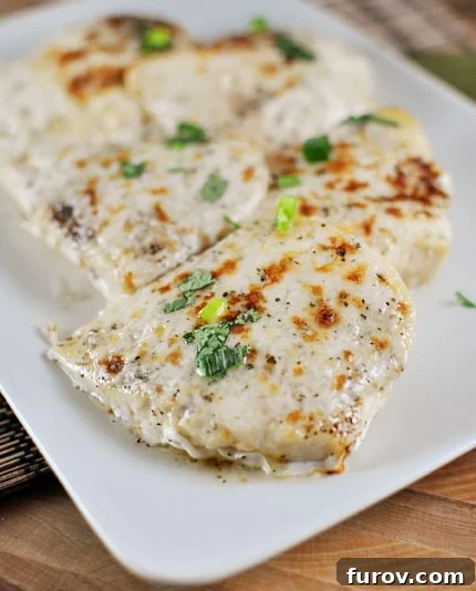 Top view of cooked chicken with melted cheese and herbs lined up on a white platter, garnished with fresh lemon slices and parsley.