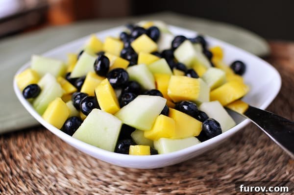 A crisp white bowl filled with a refreshing honeydew, mango, and blueberry salad, glistening with a vibrant glaze.