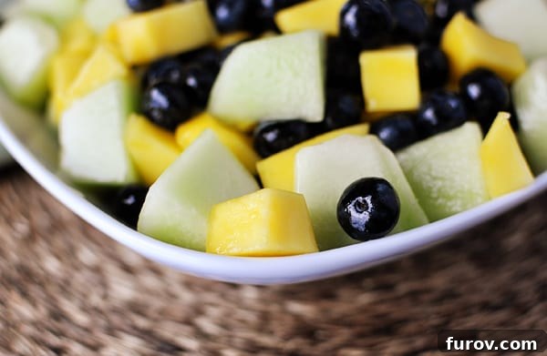 Close-up view of a vibrant honeydew, mango, and blueberry salad in a white bowl, showcasing the glistening reduction.