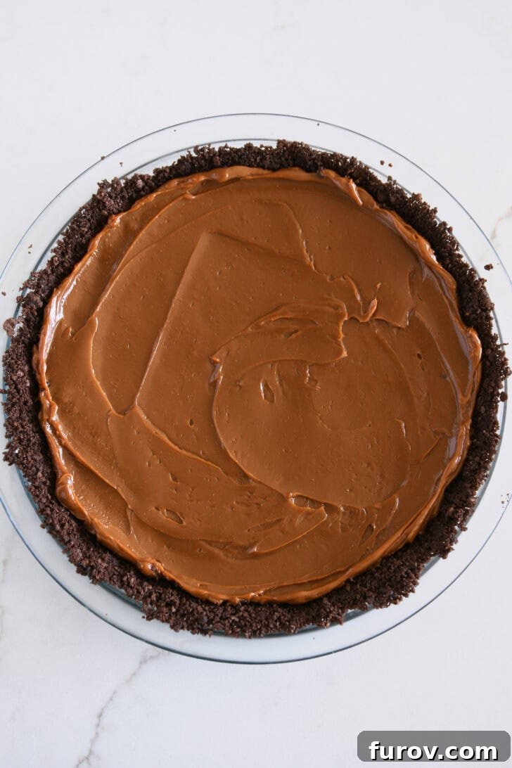 Thick, golden dulce de leche being spread smoothly over a dark chocolate cookie crust in a glass pie dish, ready for the next layer.