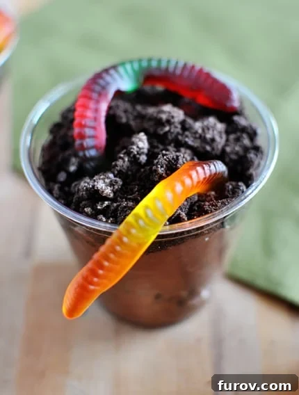 Close-up top view of a clear plastic cup filled with layers of homemade chocolate pudding and crushed Oreo cookies, topped with a vibrant gummy worm, ready to be enjoyed.