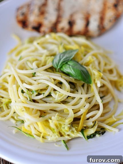 A serving of cooked spaghetti intertwined with bright green zucchini and yellow squash, topped with fresh basil leaves, with a perfectly grilled chicken breast in the background.