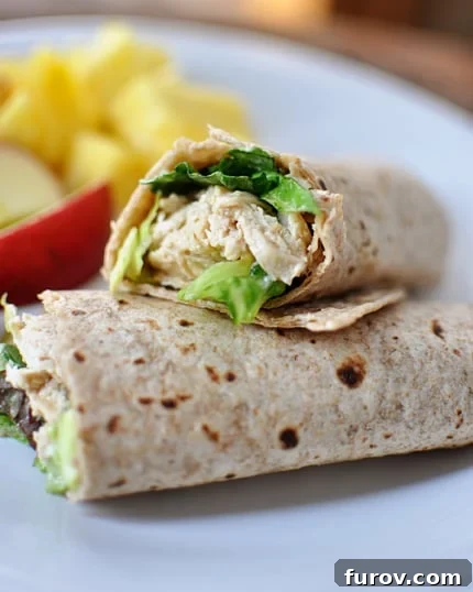 A chicken caesar salad wrap cut in half on a white plate with fresh fruit in the background, showcasing the delicious filling.