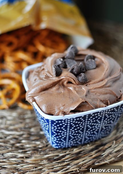 A vibrant blue ceramic bowl overflowing with a rich chocolate brownie dip, generously sprinkled with mini chocolate chips, with a rustic bag of pretzels blurring softly in the background.