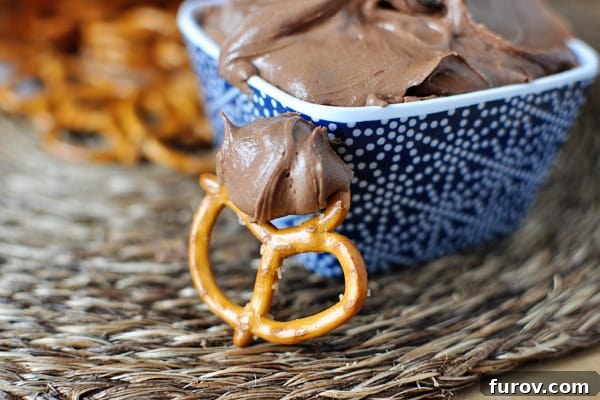 A close-up of a blue ceramic bowl featuring a smooth, rich chocolate dip. A single pretzel stick, perfectly coated with the dip, rests invitingly in front of the bowl.