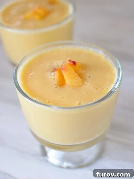 Two glass goblets filled with a vibrant peach-orange smoothie, garnished with fresh peach slices on top.