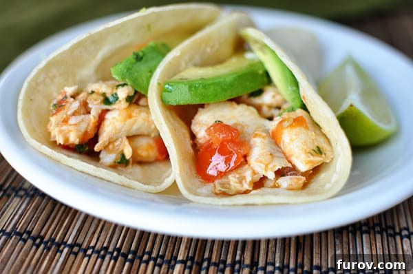 Two vibrant fish tacos in warm corn tortillas, generously garnished with fresh avocado slices and diced tomatoes, presented on a pristine white plate.