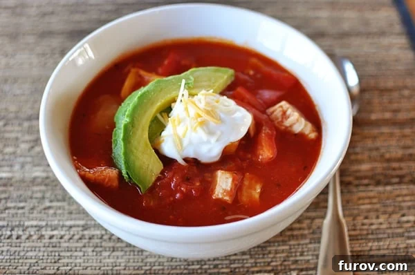 White bowl with a generous serving of red chicken chili topped with a vibrant slice of avocado and a dollop of creamy sour cream, showcasing the rich textures.